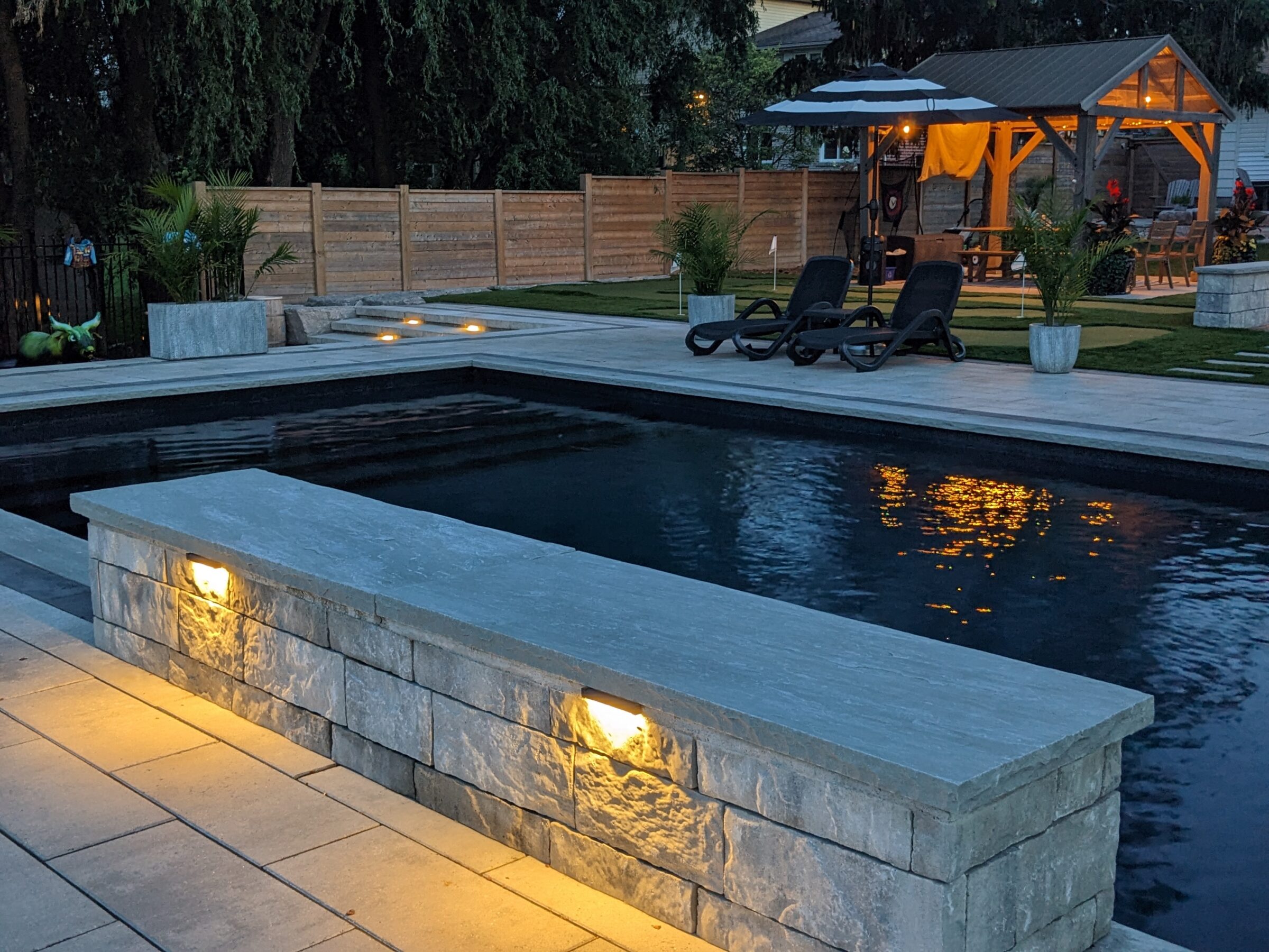 A tranquil backyard at dusk with a rectangular pool, lush greenery, illuminated gazebos, loungers, and warm lighting reflections on the water's surface.