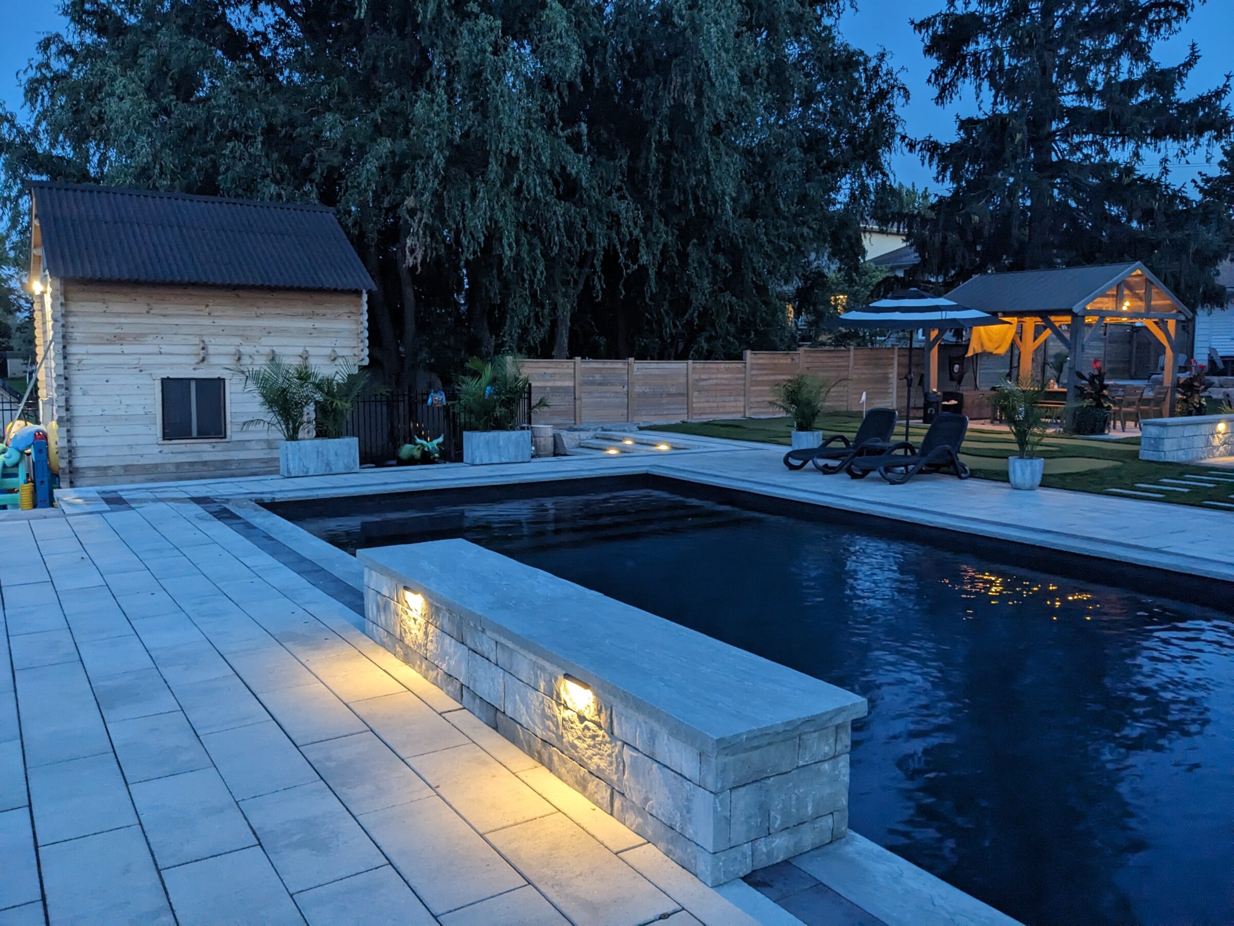 A serene backyard at dusk featuring a sleek rectangular pool, cozy gazebo, loungers, ambient lighting, wooden shed, and lush trees.
