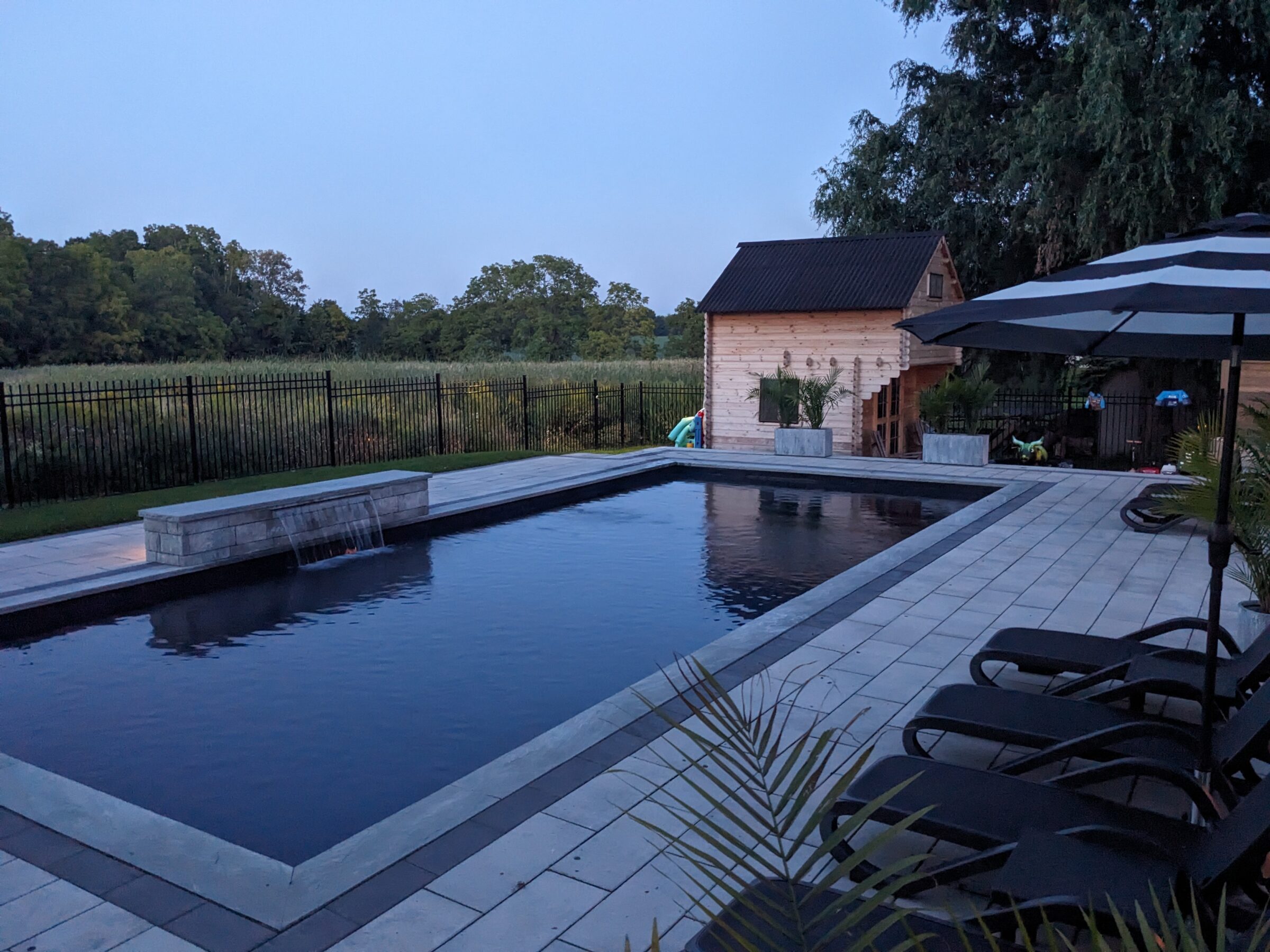 A tranquil backyard during twilight featuring a rectangular swimming pool, patio area, lounging chairs, an umbrella, and a small building with foliage.