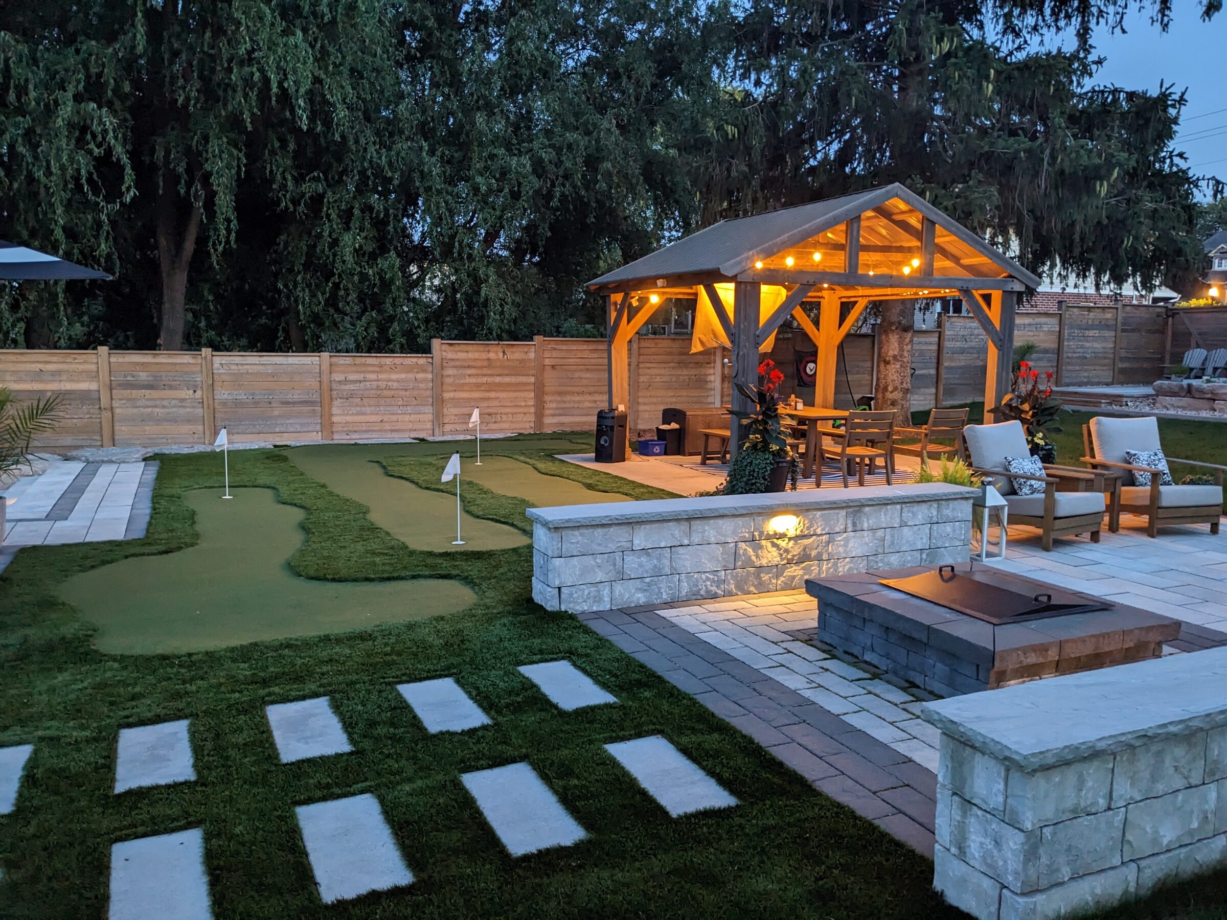 This is an outdoor living space with a mini-golf area, stone fire pit, lounge seating, and a gazebo with string lights at dusk.