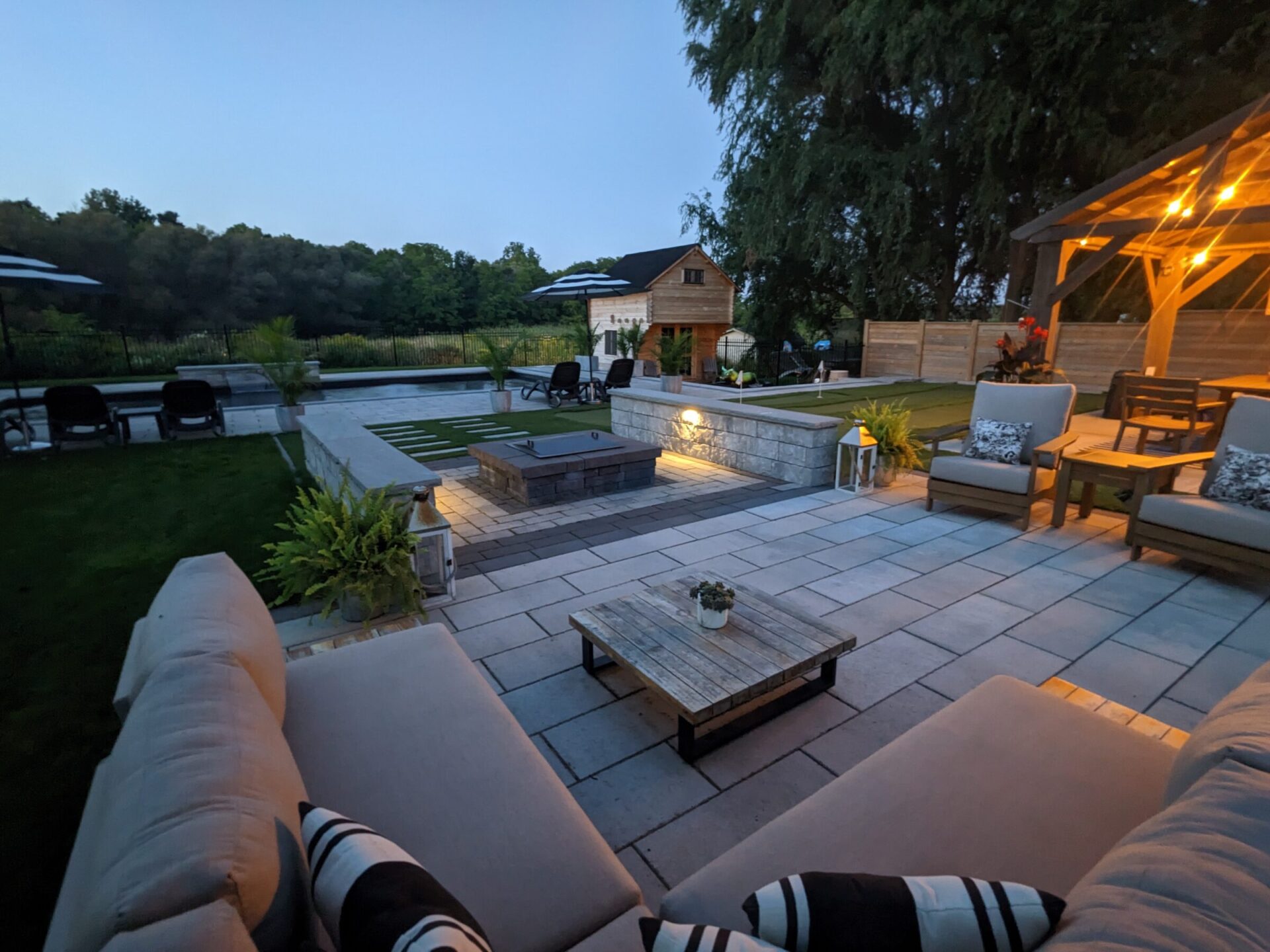 An outdoor evening setting featuring patio furniture, a fire pit, a pergola with lights, and a swimming pool, surrounded by trees and a well-kept lawn.