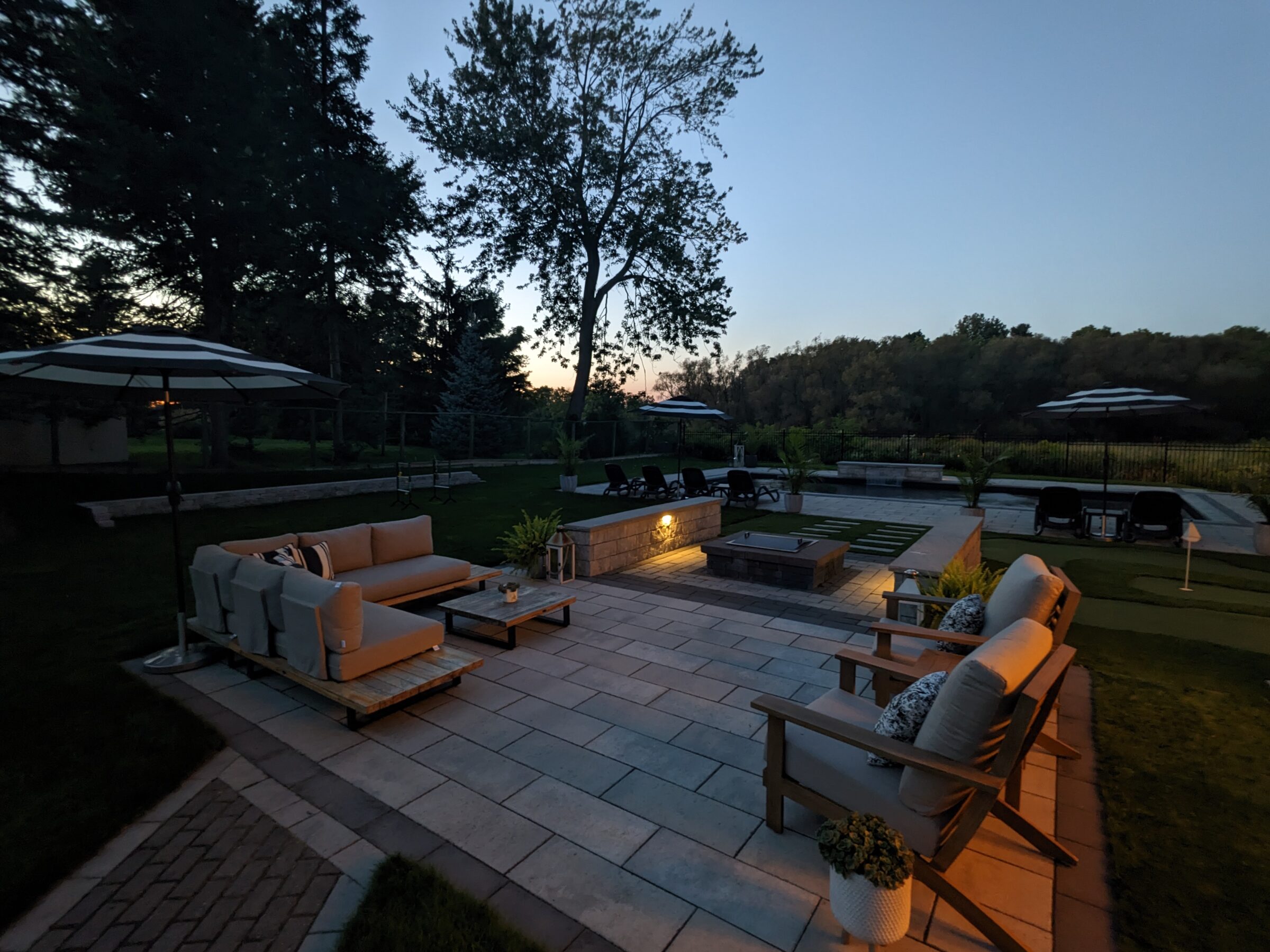 A tranquil outdoor living space at dusk featuring modern patio furniture, a fire pit, umbrellas, and a view of trees under a fading sky.